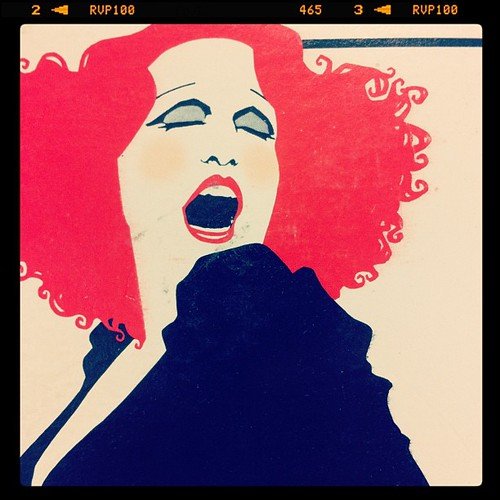 Illustration of a woman with curly red hair and an open mouth, singing or shouting, wearing a dark top.