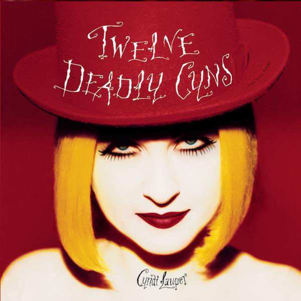 Close-up of a woman with blonde hair, pale skin, bold red lipstick, wearing a red hat that reads 'Twelve Deadly Guns'.
