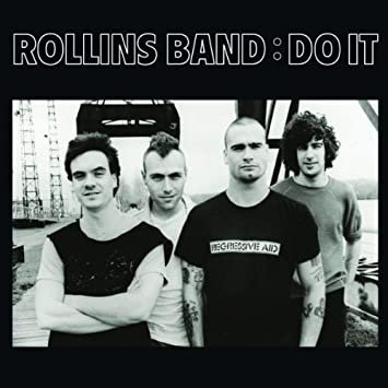 Black and white photo of the band Rollins Band with four members standing outdoors, one person in the front wearing a T-shirt, in front of an industrial backdrop.