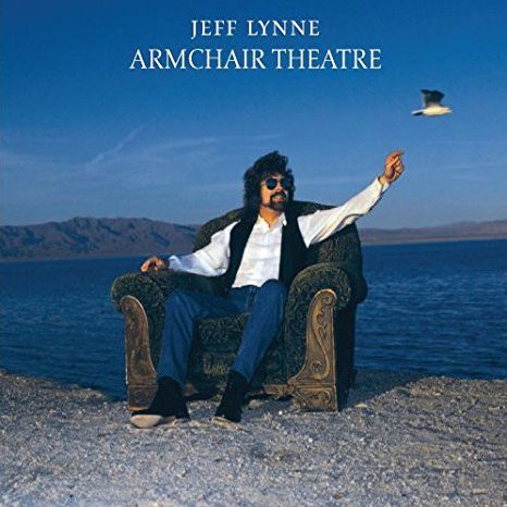 Man with sunglasses sitting on a patterned armchair on the beach, with mountains and ocean in the background, and a bird flying overhead. Text reads 'Jeff Lynne Armchair Theatre'.