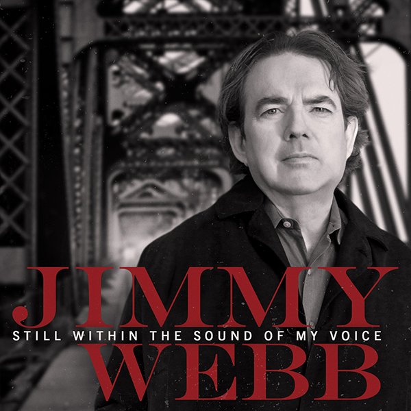 A black and white photo of Jimmy Webb with a serious expression, standing in front of a bridge. Text overlay reads: Jimmy Webb, Still Within the Sound of My Voice.