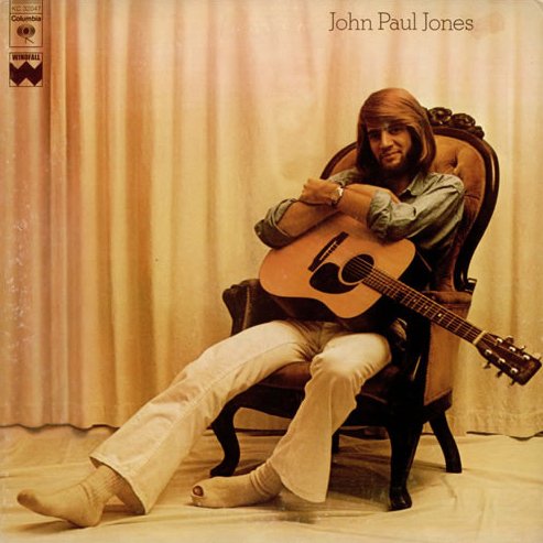 A man with long hair and a beard sitting in a vintage armchair, holding an acoustic guitar, wearing a light-colored shirt and pants, with a beige curtain background.