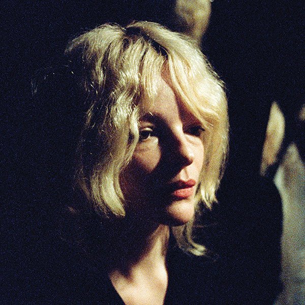 A woman with blonde, wavy hair looking slightly to the side, cast in low lighting.