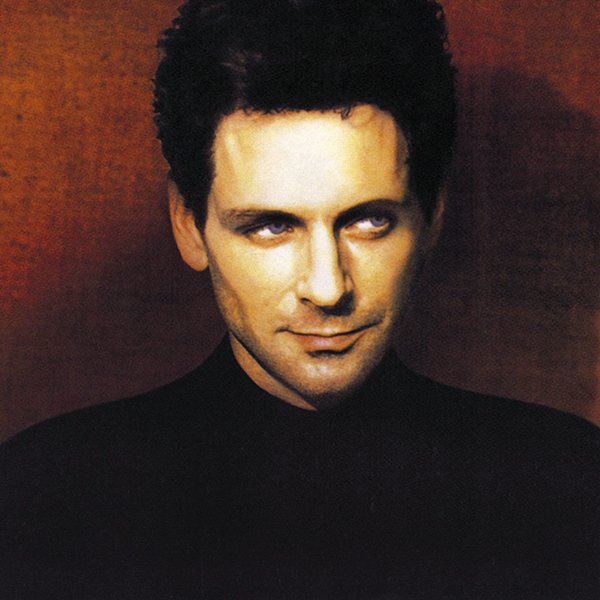 Lindsey Buckingham with dark, curly hair, wearing a black shirt, against a brown background. He is looking to the side with a slight smile.