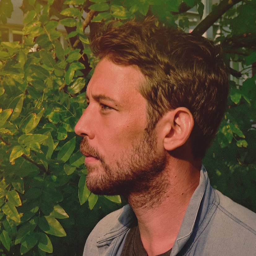 Side profile of a man with short brown hair and a beard, looking to the left, outdoors with green leafy bushes in the background.