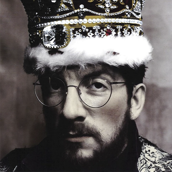A man with glasses, a beard, and a serious expression wearing a colorful, jeweled crown with a fur trim.
